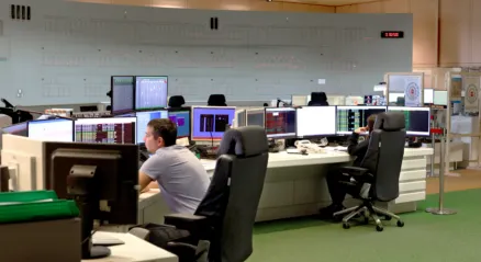 Large control centre where building services systems are monitored. A spacious room with several people working at stations equipped with multiple screens. In the background, a control panel extends from one wall to the other