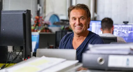 A smiling man in an office, working at a computer with several folders in front of him