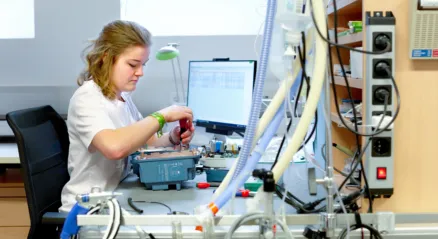 A woman in white workwear sits at a workbench, focused on assembling a small medical device. Various hoses, such as those used for ventilators, connectors and an IV drip, are attached to the workbench