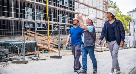 Three people wearing helmets and workwear stand in front of a construction site, showing something to one another